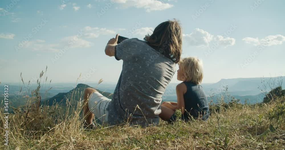Man with child boy sit on mountain top at evening takes photo on smartphone Man with child boy sit on mountain top at evening takes photo on smartphone