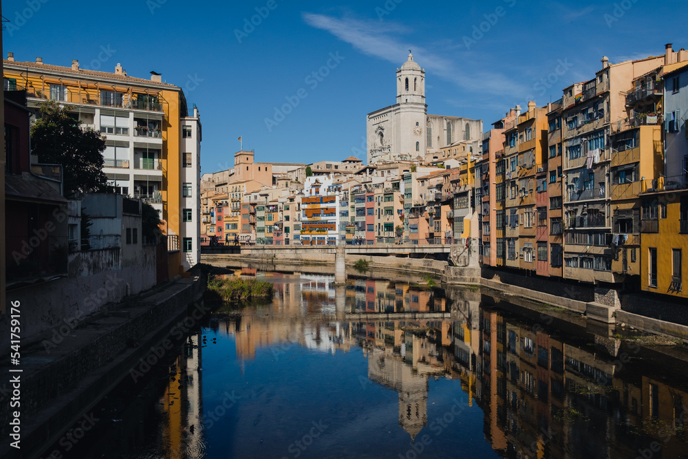 Girona, Spain on 22 October 2022: Colorful yellow and orange houses in historical jewish quarter of Girona, Catalonia, Spain reflecting on the Oñar river with clear blue sky