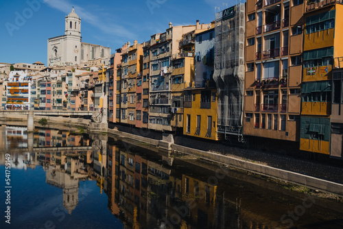 Girona, Spain on 22 October 2022: Colorful yellow and orange houses in historical jewish quarter of Girona, Catalonia, Spain reflecting on the Oñar river with clear blue sky