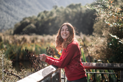blonde caucasian girl in red sweater looking at camera having fun and laughing on wooden balcony with forest and nature view, lake gunn, new zealand photo