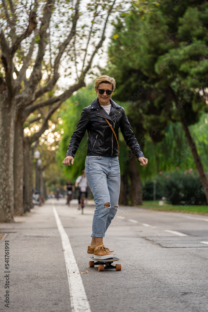 Adult woman with short hair and leather jacket learn to ride skateboard in a city. Urban lifestyle scene in Barcelona.