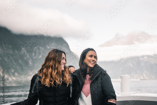 two long haired caucasian young women together looking to the side having fun and laughing on deck of tourist boat visiting fjords lake and mountain, milford sound, new zealand