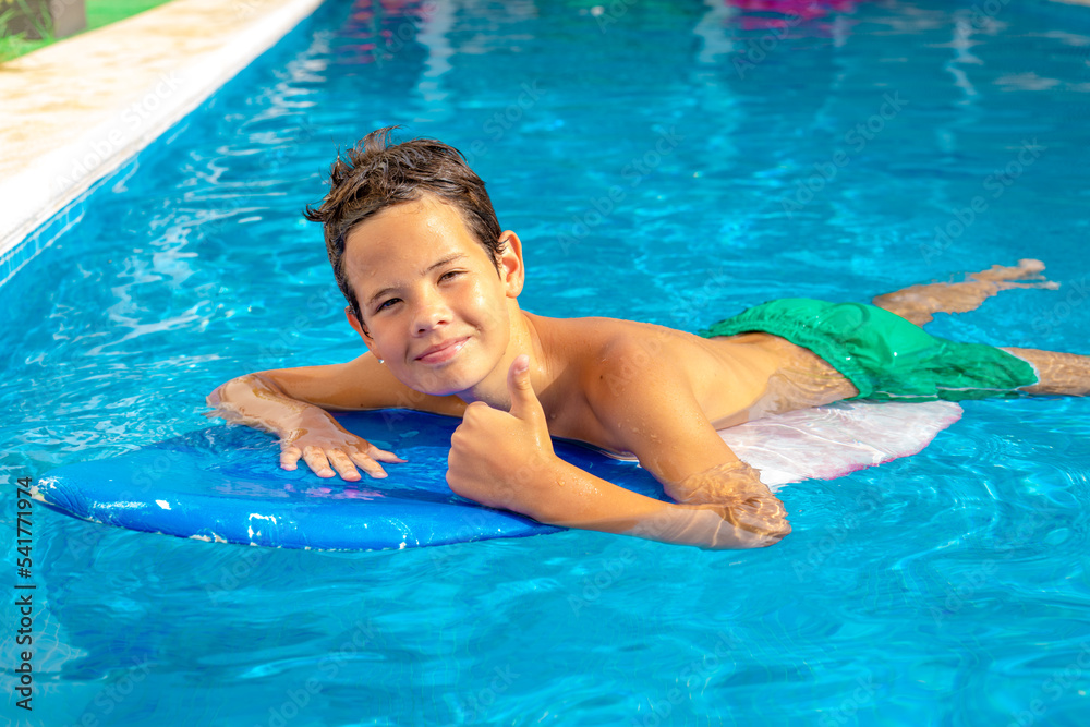 Cute smiling child boy paddling on the surf board in pool showing thumb up.