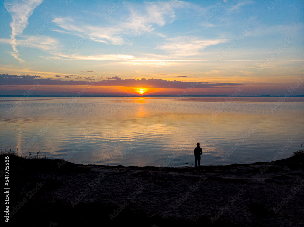drone view of a dark silhouette of a woman standing on the edge of the sea with a colourful sunset in front of her; autumn sunset over the baltic sea, hel peninsula, poland