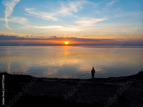 drone view of a dark silhouette of a woman standing on the edge of the sea with a colourful sunset in front of her; autumn sunset over the baltic sea, hel peninsula, poland
