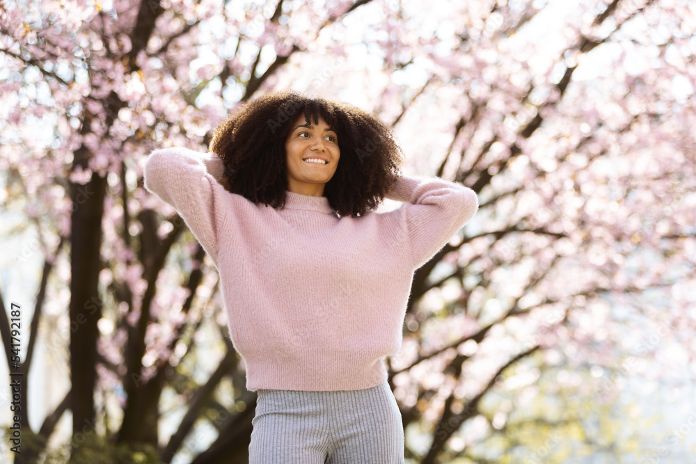 An african american woman in a pink sweatshirt with big curly hair standing in a park infront of a cherry blossom tree. An african american woman in a pink sweatshirt with big curly hair standing in a park infront of a cherry blossom tree.