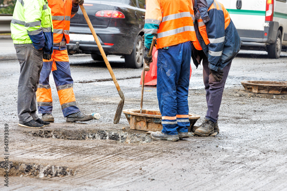 Road workers in orange overalls are repairing old sewer manholes on the roadway against the backdrop of passing cars. Copy space. Road workers in orange overalls are repairing old sewer manholes on the roadway against the backdrop of passing cars. Copy space.