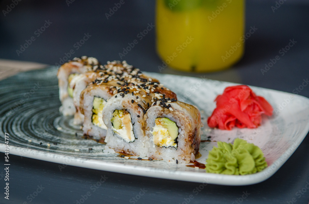 Appetizing portion of sushi rolls philadelphia with eel, with soft cheese, cucumbers, avocado and ginger with wasabi, on a gray plate on a black table, on the background of yellow fruit juice Appetizing portion of sushi rolls philadelphia with eel, with soft cheese, cucumbers, avocado and ginger with wasabi, on a gray plate on a black table, on the background of yellow fruit juice