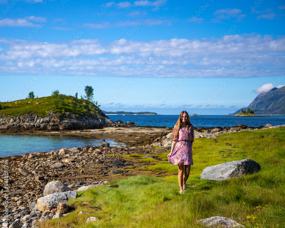a long-haired girl in a colourful dress walks by the sea on the island of senja in norway; green grass and colourful houses by the sea under the mighty mountains