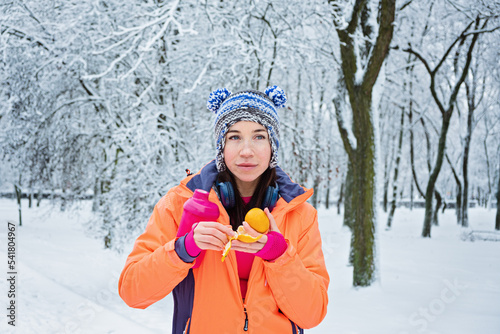 Winter Run Fuel. Sporty fitness woman in headphones eating tangerine and drinking water outdoors in winter snowy park forest. Food and water for jogging in winter