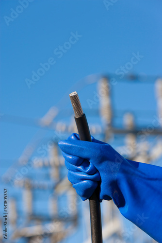 A gloved hand holds wires in front of an electrical utility substation.