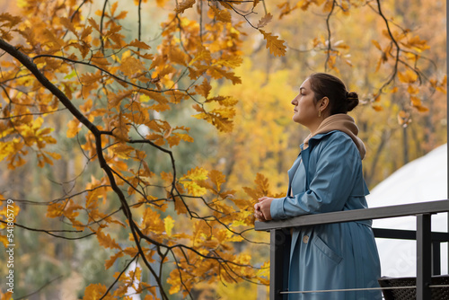 Woman leans on balcony railing with hands breathing deeply and smiling lightly. Female stands near tree with bright yellow leaves enjoying autumn nature, copyspace