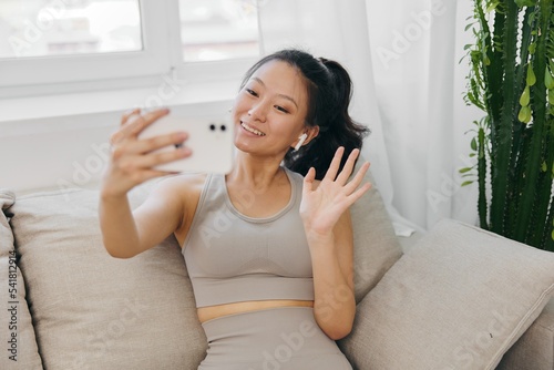 Athletic Asian woman in yoga costume blogger films herself on her phone camera sitting on the couch and smiling with headphones, freelance work from home, recording sports workout lifestyle body care