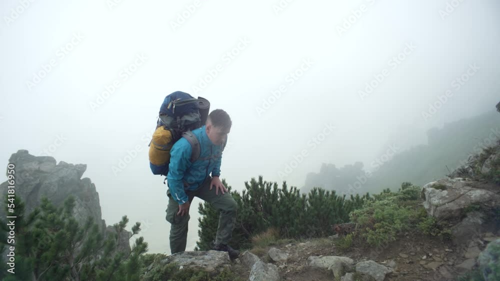 A man with a backpack climbs rocky mountains into the fog. A tourist conquers a mountain peak in the rain. A boy walks uphill next to large stones in the Carpathians. Concept: submission, fatigue. 4k