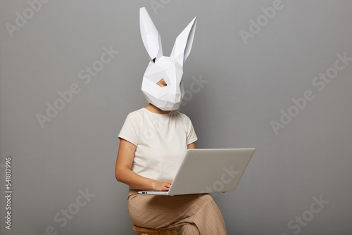 Image of unknown female wearing bunny mask and white t-shirt isolated over gray background, sitting on chair and working on personal computer, doing her work.