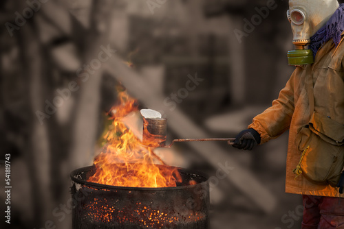 The life of a survivor of a man-made disaster. A man in a gas mask holds a tin can over a rusty barrel with a flame. Selective focus.