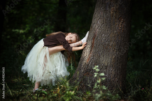 Pretty little slim girl doing stretching exercises as ballet dancer ballerina near big tree in summer forest or park, with leg raised up, dressed in fluffy dress and ballet shoes, dark background