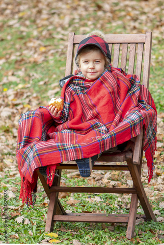 Portrait of a little girl 4-5 years old in a plaid and a cap, eats a bun, sits in a wooden chair on a blurred background of nature.