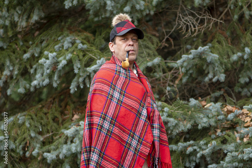 Portrait of an elderly man 45-50 years old with a smoking pipe in his mouth  with a plaid on his shoulders and a stylish cap against the background of fir trees.