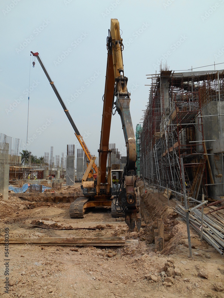 SELANGOR, MALAYSIA –MARCH 15, 2016: Sheet pile cofferdam driven machine at the construction site. The machine drove the sheet pile to the earth using vibrated hydraulic arm. Workers control. SELANGOR, MALAYSIA –MARCH 15, 2016: Sheet pile cofferdam driven machine at the construction site. The machine drove the sheet pile to the earth using vibrated hydraulic arm. Workers control.
