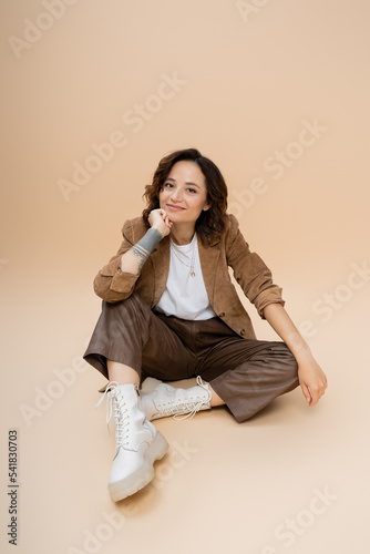 brunette woman with wavy hair wearing trendy autumn clothes while sitting with hand near face on beige background