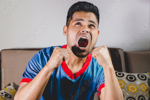 Joven fanático del fútbol, gritando gol, apoyando a su equipo sentado en el sofá de casa viendo la transmisión en vivo del encuentro. Concepto de mundial de futbol e hinchas del futbol en el mundo.