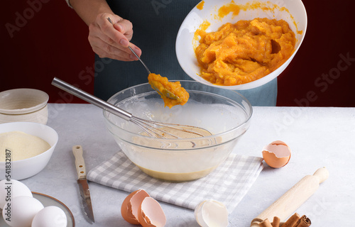 Woman add a pumpkin puree into a bowl with filling for pumpkin pie for mix with whisk on a gray table surrounded by ingredients in the kitchen. Home cooking concept, traditional pie recipe