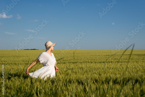 Young woman in white dress and hat walking in the wheat field on a sunny spring day