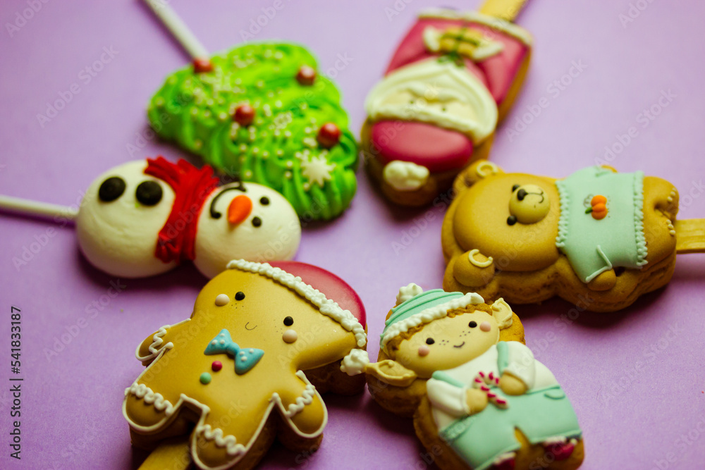 Cookies on sticks in a shape of Santa Claus, elf character, Gingerbread man, snowman, bear, New Year 2023 tree, lollipop on purple background top view. Cute treats, sweets, candies for Christmas fun.