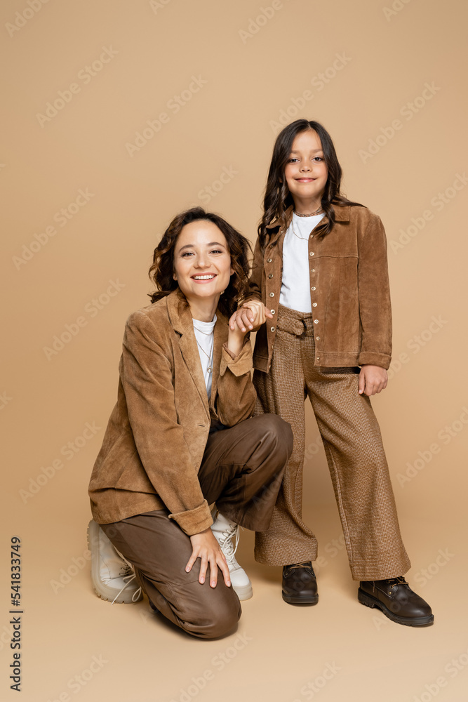 Fototapeta premium smiling mother and child in suede jackets and trendy clothes holding hands on beige background