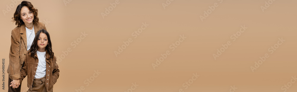 girl in trendy suede jacket posing with hand in pocket near cheerful mom isolated on beige, banner