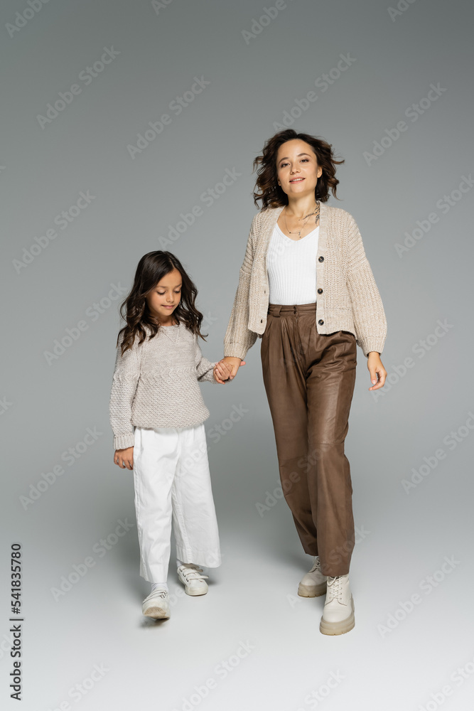 trendy woman smiling at camera while holding hands with daughter and walking on grey background trendy woman smiling at camera while holding hands with daughter and walking on grey background