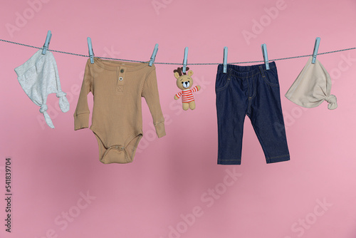 Different baby clothes and bear toy drying on laundry line against pink background