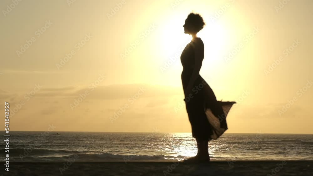 Time lapse of a silhouette of a woman in a midi dress, standing sideways to the camera against the backdrop of a sunset and seashore. The sun creates a rainbow aura around the woman.