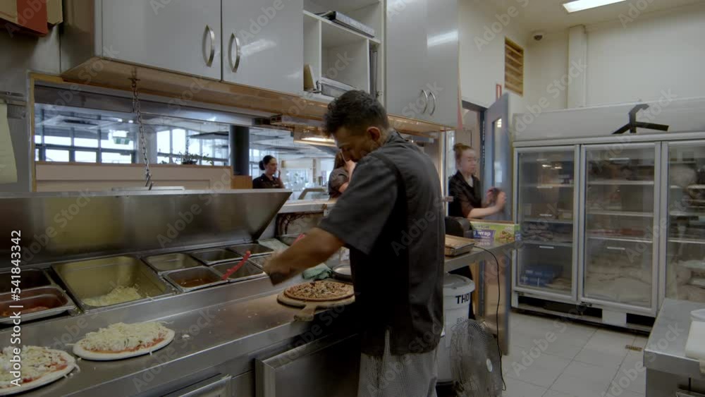 Kitchen worker cuts pizza fresh out of the oven, then grabs plate. Inside commercial kitchen. Kitchen worker cuts pizza fresh out of the oven, then grabs plate. Inside commercial kitchen.