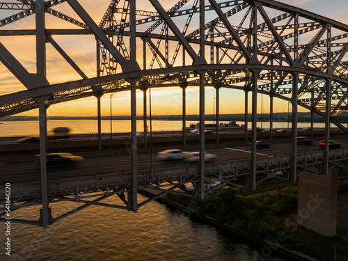 A large truss bridge over a vast body of water is seen at sunset as cars, motion blurred, travel across the multi-lane highway.