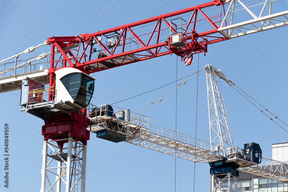 A close-up of two large, modern construction cranes, one red and one blue, are seen on a clear, blue sky day. A close-up of two large, modern construction cranes, one red and one blue, are seen on a clear, blue sky day.