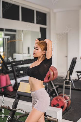 A sexy and sultry asian woman in a black crop top looking at camera while doing tricep stretches before a workout session at the gym.