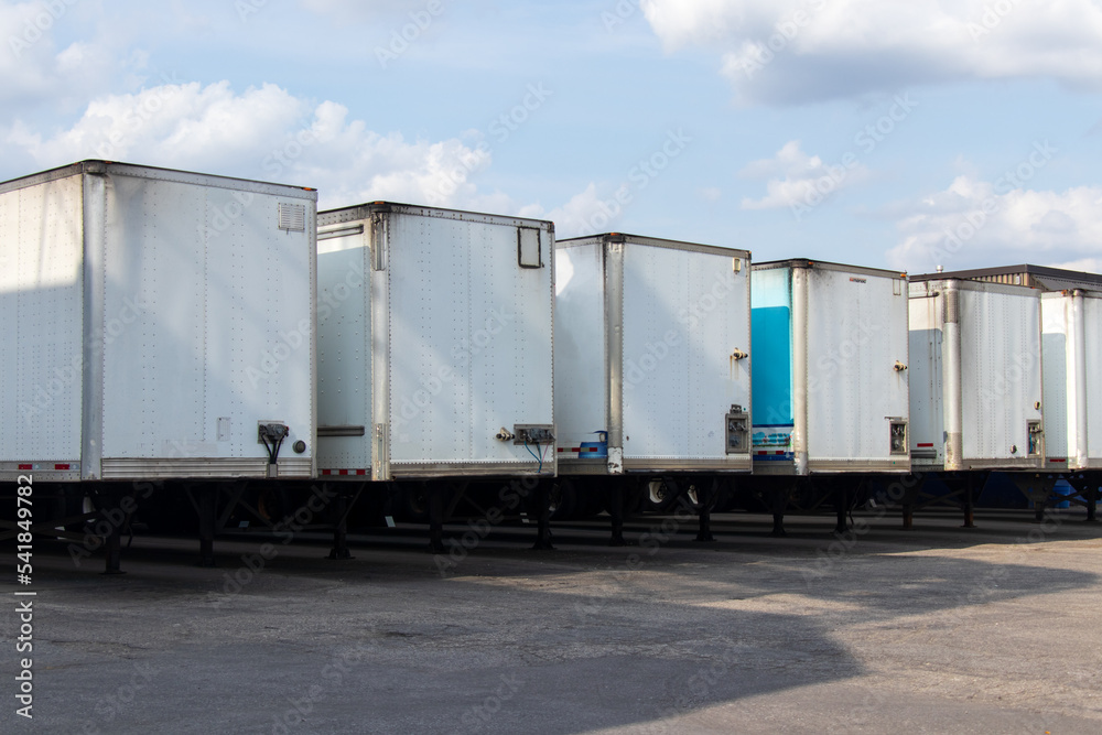 A row of white transport trailers are lined up outside a warehouse on a partly cloudy, sunny day. A row of white transport trailers are lined up outside a warehouse on a partly cloudy, sunny day.
