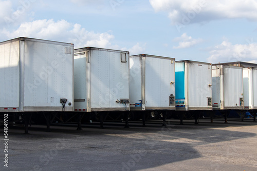 A row of white transport trailers are lined up outside a warehouse on a partly cloudy, sunny day.