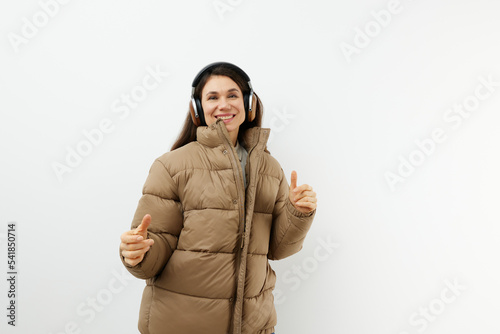 beautiful happy brunette listens to music in brown headphones while standing in a warm winter stylish jacket and smiles sweetly with enjoyment. Horizontal studio photo on a light background