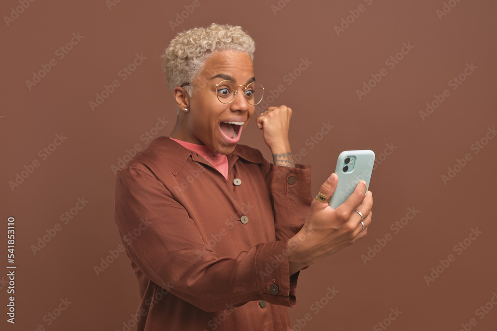 Fototapeta premium Young amazed African American woman shouting and making YES gesture looking at screen of mobile phone rejoicing at victory of sports team or jackpot in online casino, stands on brown background