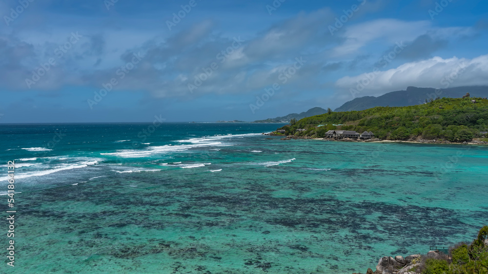 Beautiful tropical seascape. Waves of turquoise ocean foam over coral reefs. On the shore, overgrown with tropical vegetation, the houses of the hotel are visible. Blue sky with clouds. Seychelles