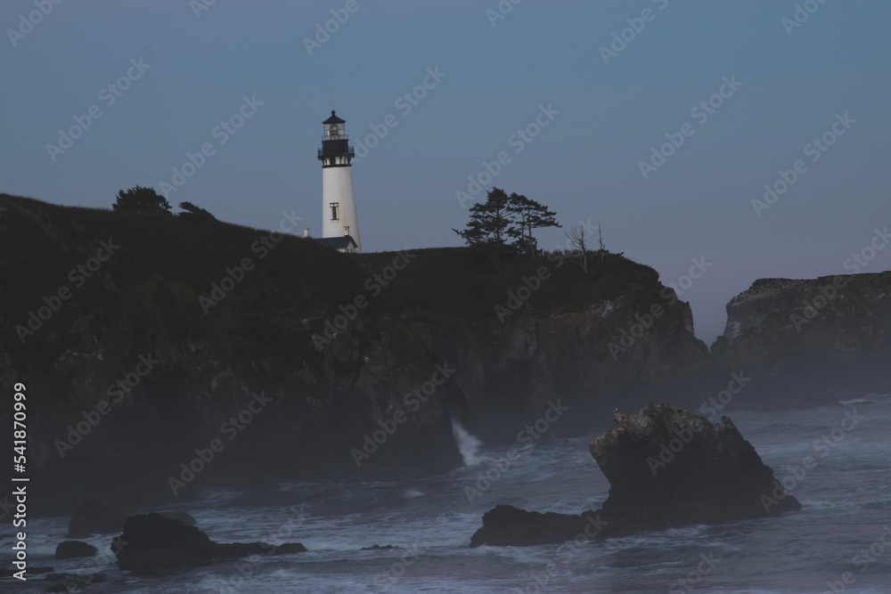 Sunrise at the Yaquina Head Lighthouse in Newport, Oregon.  The lighthouse is on a rocky outcropping, with the sea below with a layer of fog on the beach. 