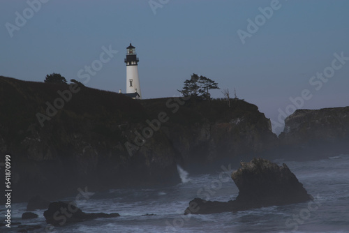 Sunrise at the Yaquina Head Lighthouse in Newport, Oregon. The lighthouse is on a rocky outcropping, with the sea below with a layer of fog on the beach. 