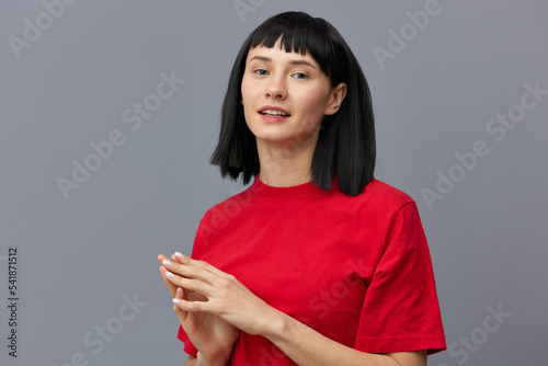 a sweet, modest, attractive woman stands on a gray background in a red T-shirt and smiling pleasantly at the camera keeps her fingers folded in a relaxed pose