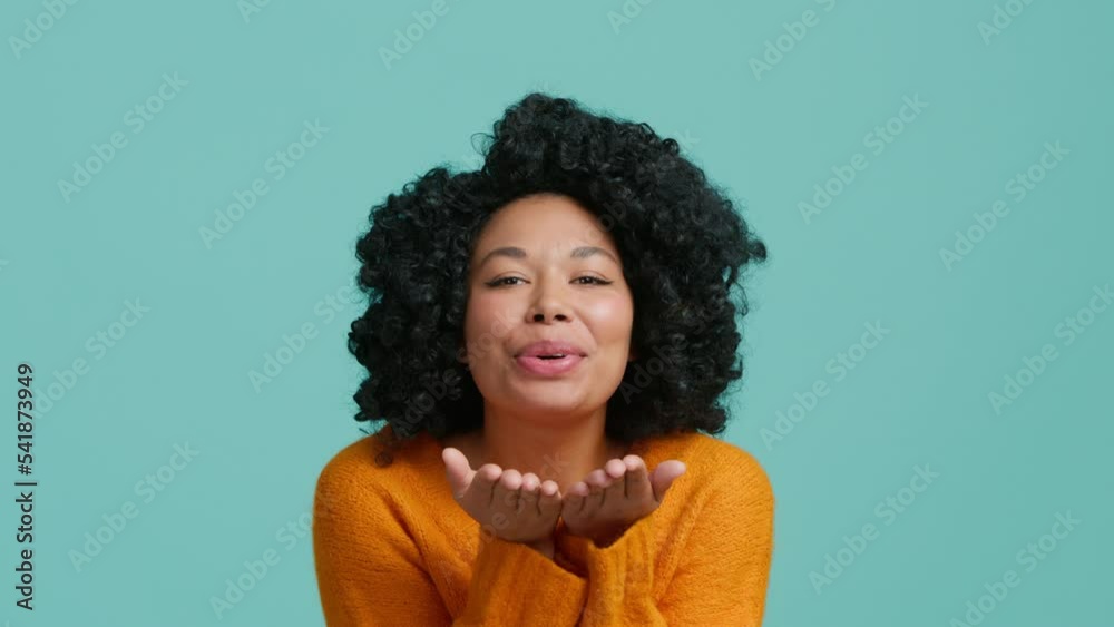 Cute smiling African American lady in bright orange sweater send air kiss from both palm of hands to boyfriend, romantic gesture of true love, friendship and passion isolated pastel blue background Cute smiling African American lady in bright orange sweater send air kiss from both palm of hands to boyfriend, romantic gesture of true love, friendship and passion isolated pastel blue background