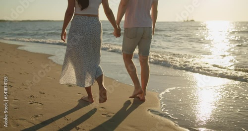 Young loving couple spend time on the beach at sunset. Man and woman enjoying relaxing walk on the beach holding hands. Only legs are taken into the frame. Travel vacation lifestyle.