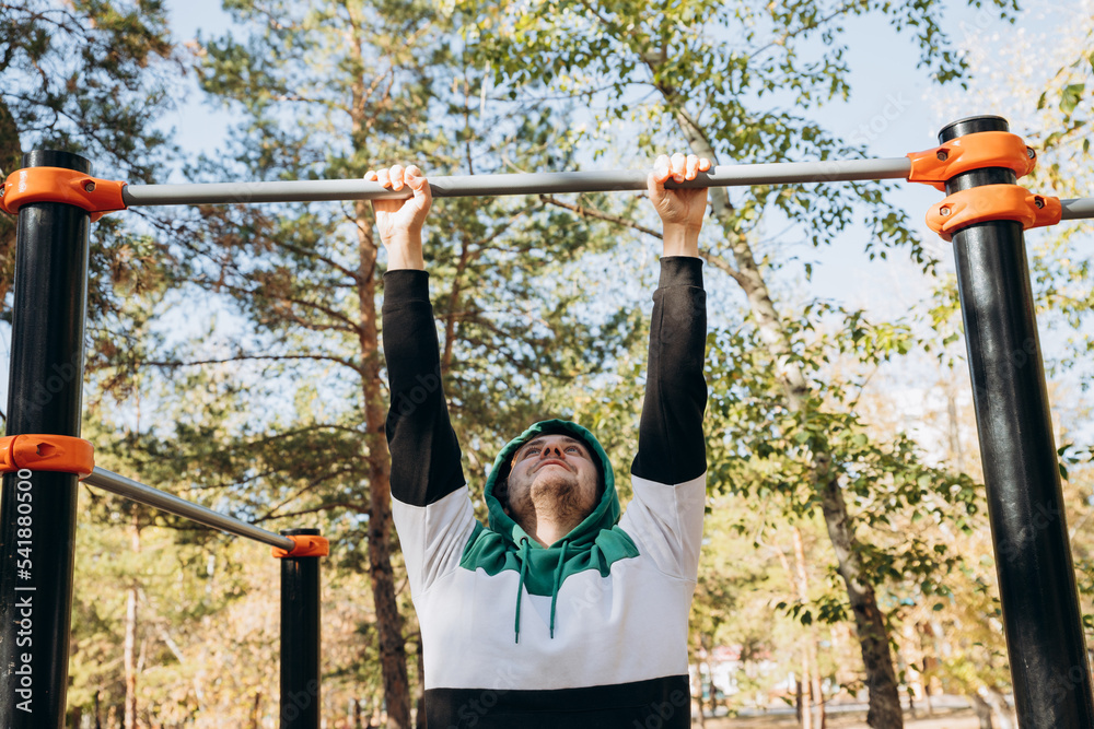 Fototapeta premium Young caucasian unshaven man 34 years old doing pull ups at the gym, unsporting non- athletic man hanging on bars on sunny autumn day. Outdoor park sports equipment. Lifestyle portrait.