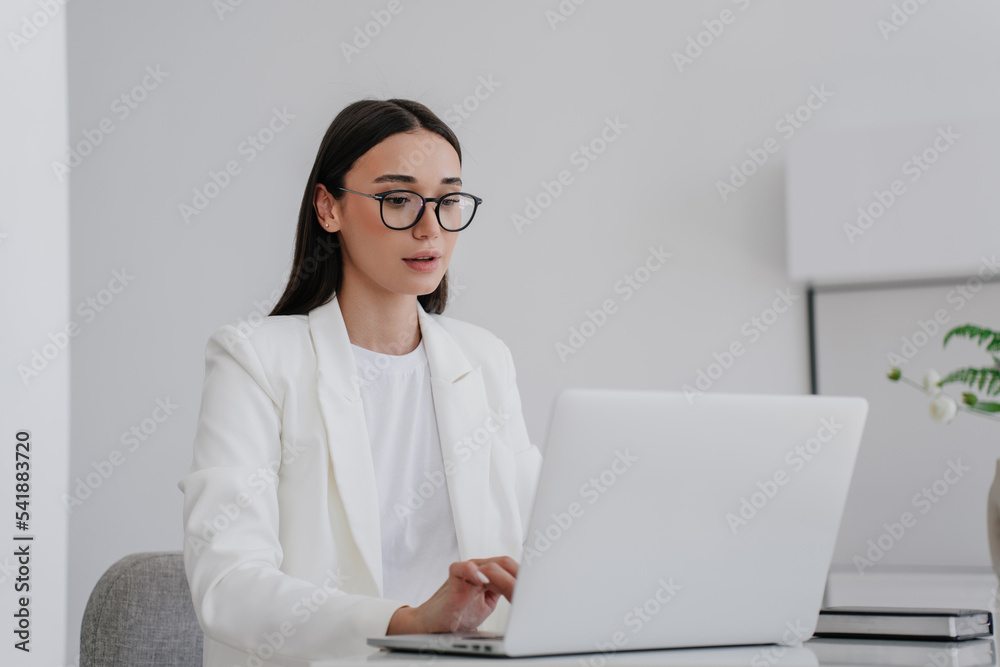 Elegant brunette young businesswoman remote working home, in white suit, using laptop sitting at desk in glasses. Attractive student girl having distant lesson. Mockup, Business and technologies. Elegant brunette young businesswoman remote working home, in white suit, using laptop sitting at desk in glasses. Attractive student girl having distant lesson. Mockup, Business and technologies.
